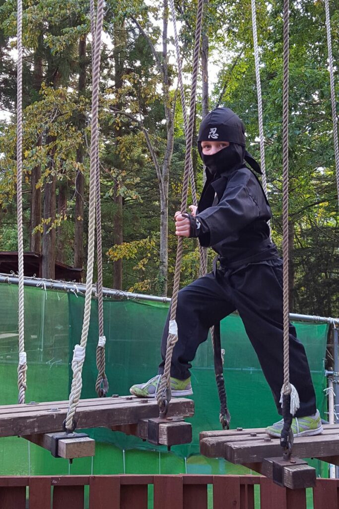 A child dressed in a black ninja outfit crosses a suspended wooden plank bridge while holding onto ropes. The child appears focused and determined, with a forested area visible in the background.