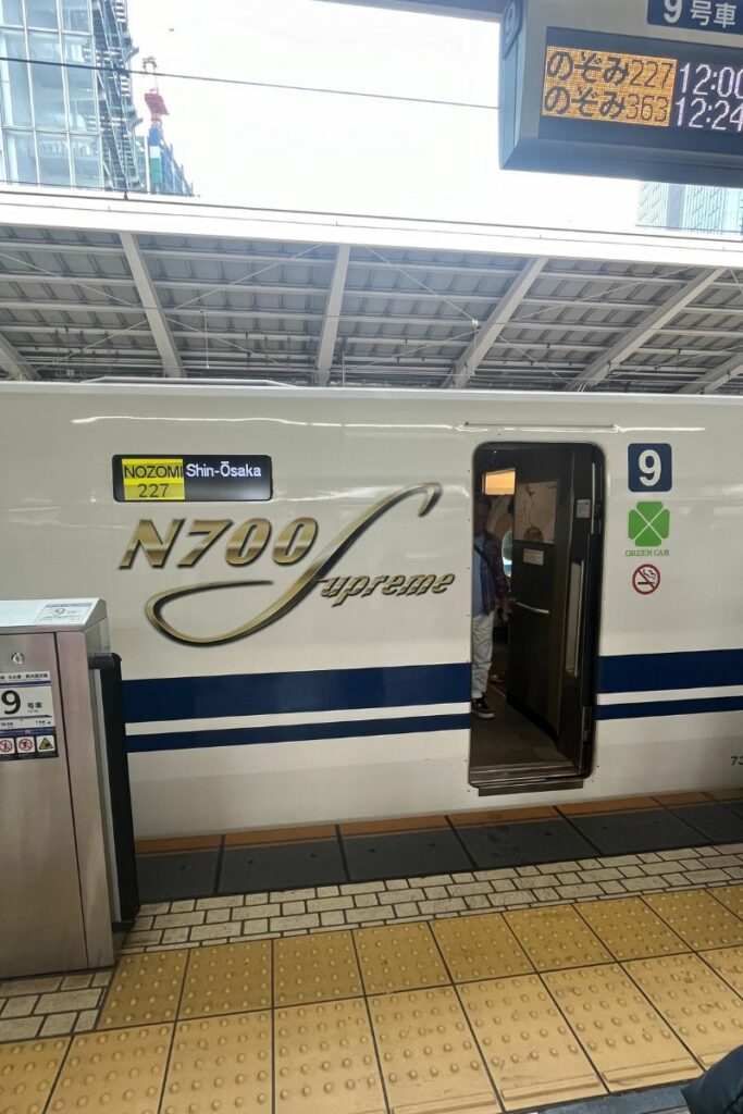 A N700 Supreme Shinkansen train is stationed at a platform, with the door to car number 9 open for boarding. The exterior of the train features the "N700 Supreme" logo and signage indicating it is a "Nozomi" service to Shin-Osaka. The green icon next to the door denotes that this car is a Green Car (business class), and there are signs for no smoking and emergency exit instructions. The platform has tactile paving for accessibility.