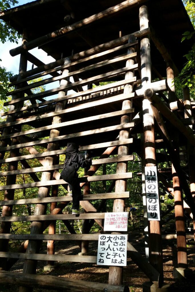A child dressed in a black ninja outfit climbs a large wooden structure made of horizontal logs. The structure has signs with Japanese text, and the background features a forested area with sunlight filtering through the trees.
