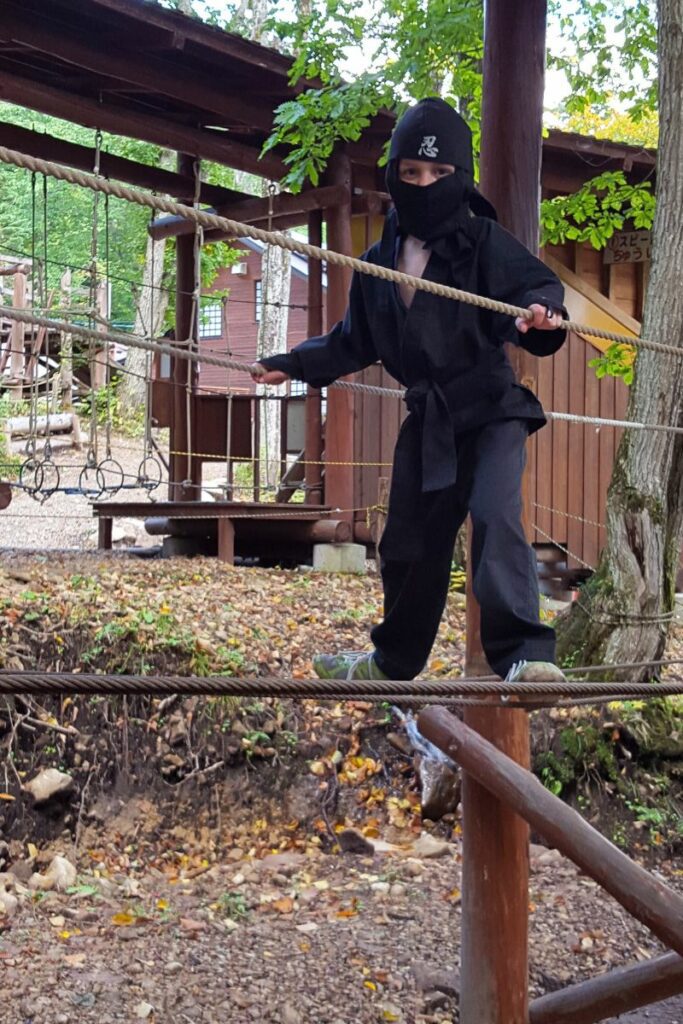 A child dressed in a black ninja outfit carefully traverses a rope bridge, balancing with one foot on a lower rope and holding onto an upper rope for support. The background shows a wooded area and part of an obstacle course.
