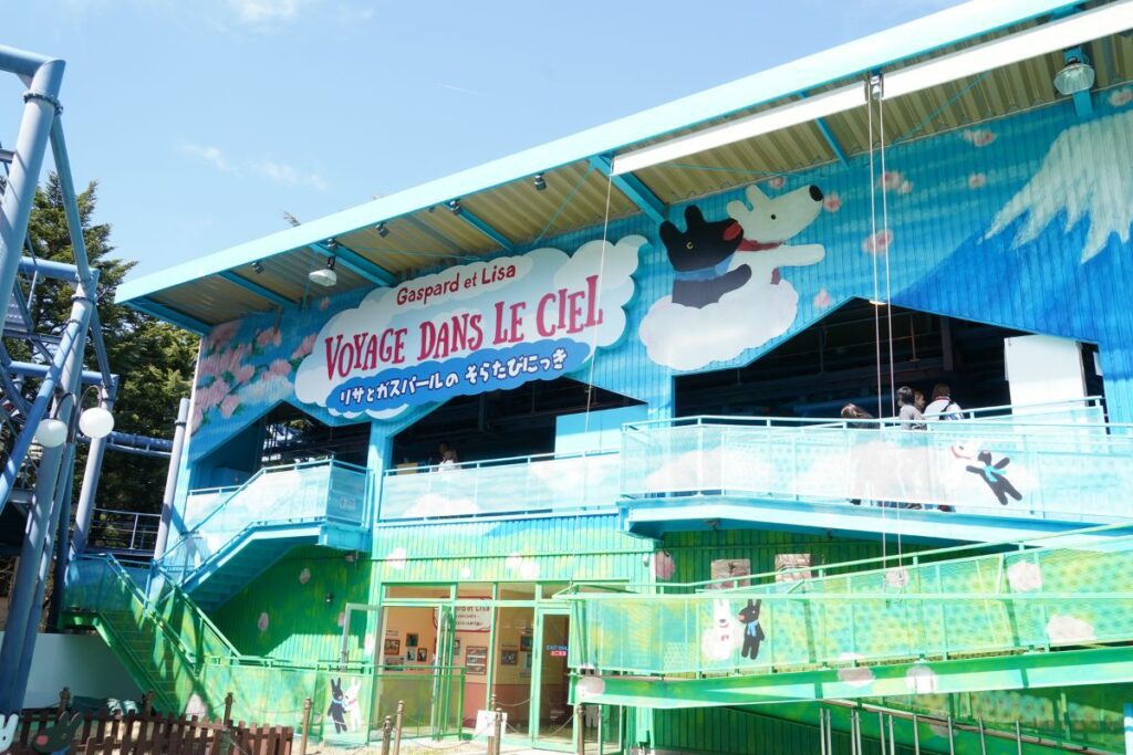 Entrance to the 'Voyage Dans Le Ciel' ride at Fuji-Q Highland, themed around the characters Gaspard and Lisa. The ride's facade is adorned with vibrant blue and cloud imagery, with illustrations of the characters floating on clouds. The entrance features multiple levels with transparent blue walkways, enhancing the airy and whimsical theme of the attraction.