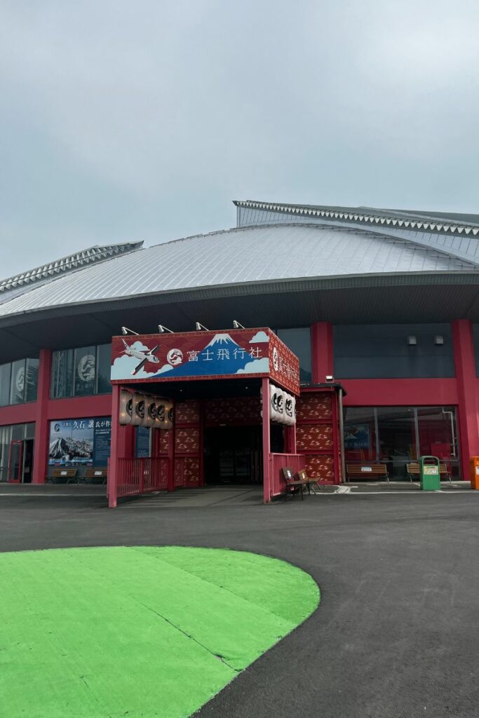 The entrance to Fuji Airways at Fuji-Q Highland, styled with a vibrant red and blue traditional Chinese architectural theme. The facade features intricate designs and Japanese characters, contrasting with the modern grey metal roof of the larger building behind it. In front of the entrance, there's a green decorative patch on the ground, adding a playful touch to the setting.