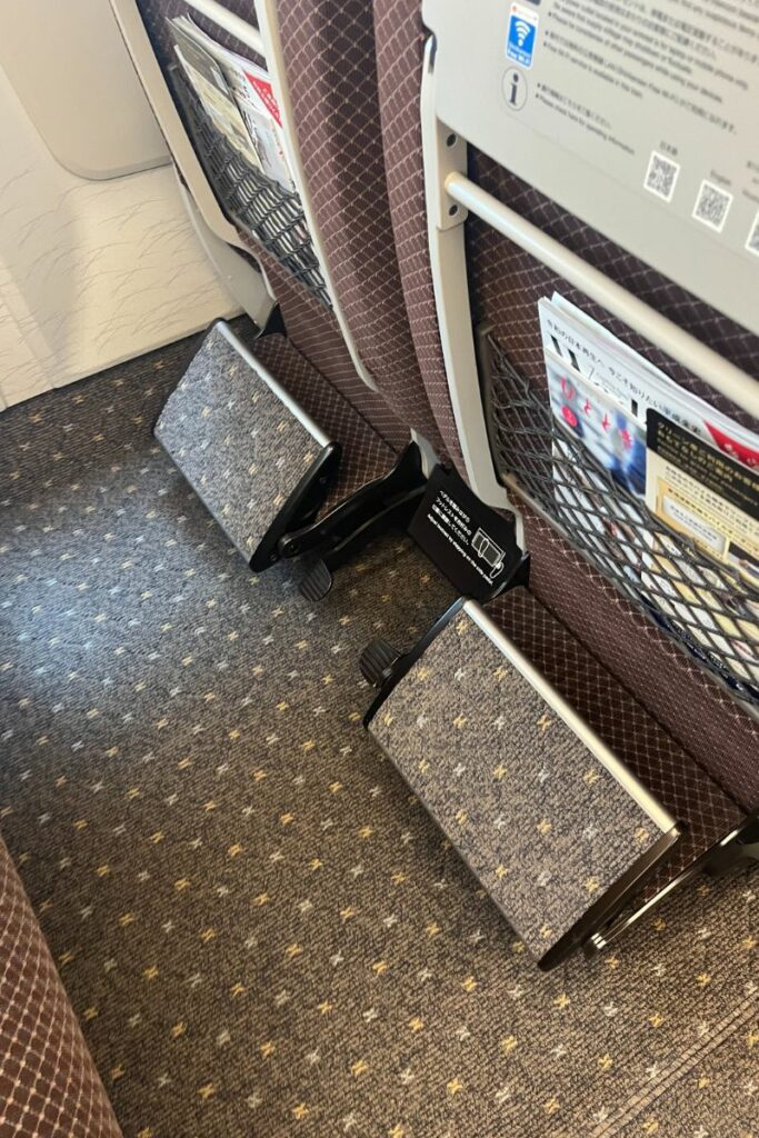 The footrests in the Green Car (first-class) of an N700 Shinkansen train are shown, positioned at the back of the seats in front. The footrests are adjustable and covered in a patterned fabric matching the seat upholstery. There are also mesh pockets on the seatbacks containing informational materials for passengers. The floor has a patterned carpet, providing a comfortable and premium travel experience.