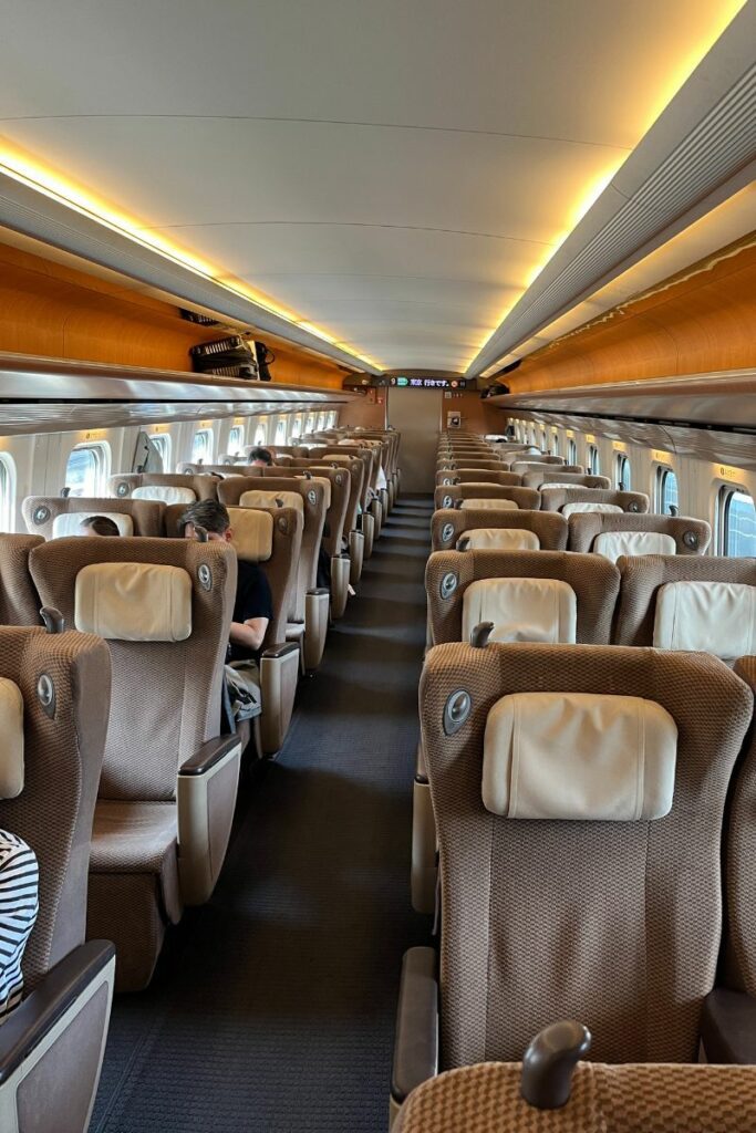 Interior of the Green Class cabin on the E5 Shinkansen, featuring spacious seating arranged four across in a 2-2 configuration. The seats are upholstered in textured brown fabric with adjustable headrests and ample legroom, illuminated by soft overhead lighting. The aisle is clear, providing a comfortable and quiet travel environment.