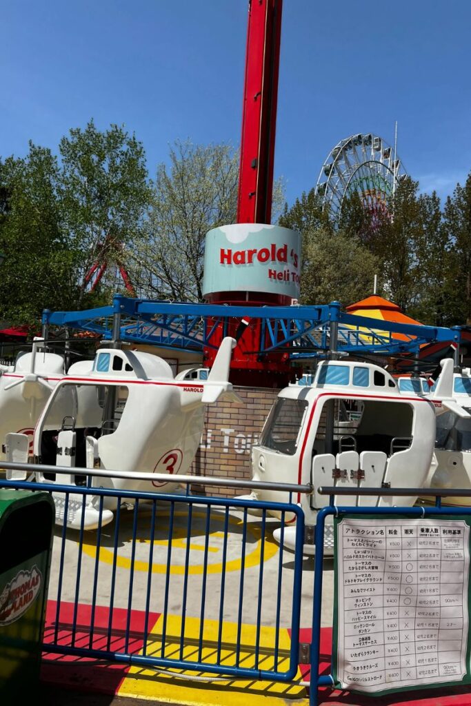 Harold's Heli Tour ride at an amusement park, featuring a vertical central column with several small helicopter-shaped cars attached around it, ready to be lifted into the air. The helicopters are painted white and labeled 'Harold', set against a vibrant backdrop that includes other amusement rides like a Ferris wheel. A clear blue sky enhances the colorful and lively setting.
