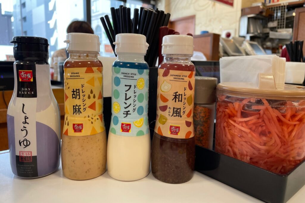 A selection of condiments at a Japanese gyudon restaurant, featuring bottles of soy sauce, sesame dressing, French-style dressing, and Japanese-style dressing, alongside a container of beni-shoga (pickled red ginger). The condiments are neatly arranged with chopsticks and napkins in the background.
