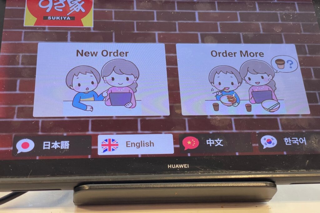 A digital ordering screen at a Sukiya restaurant, showing options for "New Order" and "Order More" with cartoon illustrations of people placing orders. Below these options, there are language selection buttons for Japanese, English, Chinese, and Korean. The screen is set against a brick wall background.