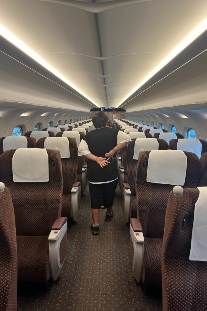 The interior of a Green Car (first-class) on an N700 Shinkansen train features rows of spacious, brown upholstered seats with white headrest covers. A passenger is walking down the aisle, and the overhead bins are empty, providing a clean and organized appearance. The car is well-lit with a modern design and large windows along the sides.