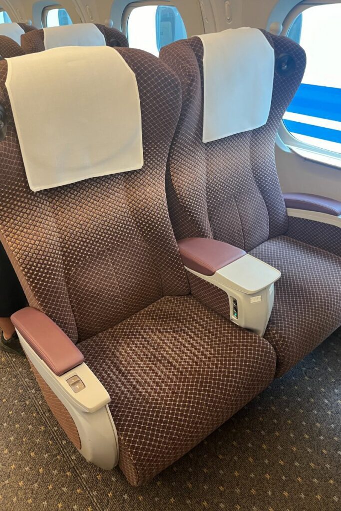 A close-up view of two plush seats in the Green Car (first-class) of an N700 Shinkansen train. The seats are upholstered in brown fabric with a diamond pattern and feature white headrest covers and wide, cushioned armrests. The seats are positioned next to a large window, providing a comfortable and spacious seating arrangement for passengers.