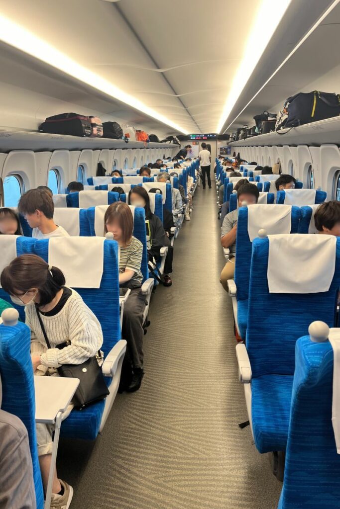 The interior of an N700 Shinkansen train's ordinary class car is shown, with passengers seated in rows of blue seats. Overhead compartments are filled with luggage, and passengers are engaged in various activities like reading and using electronic devices. The car has a bright and modern design, with a center aisle and windows along both sides.