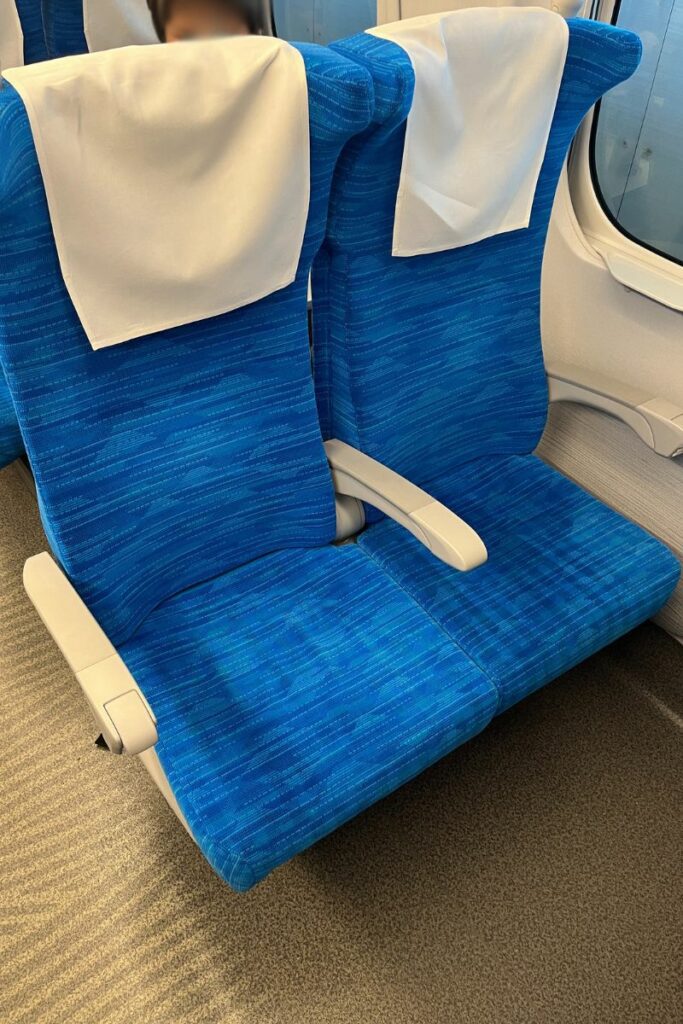 A close-up view of two blue seats in the ordinary class section of an N700 Shinkansen train. The seats have white headrest covers and are positioned next to a window, offering a clean and comfortable appearance with ample legroom. The armrest between the seats can be seen, and the floor has a patterned carpet.