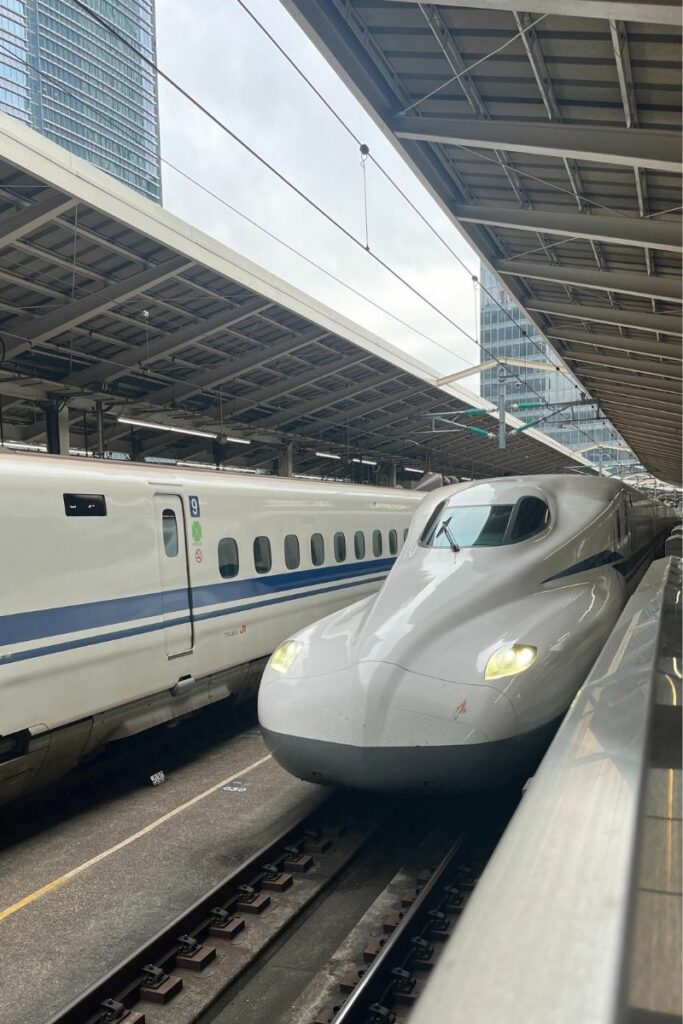 A sleek N700 Shinkansen bullet train is stationed on the tracks at a covered platform in Tokyo. The train's aerodynamic design and modern features are highlighted as it waits next to another Shinkansen train. Tall buildings can be seen in the background, emphasizing the urban setting.