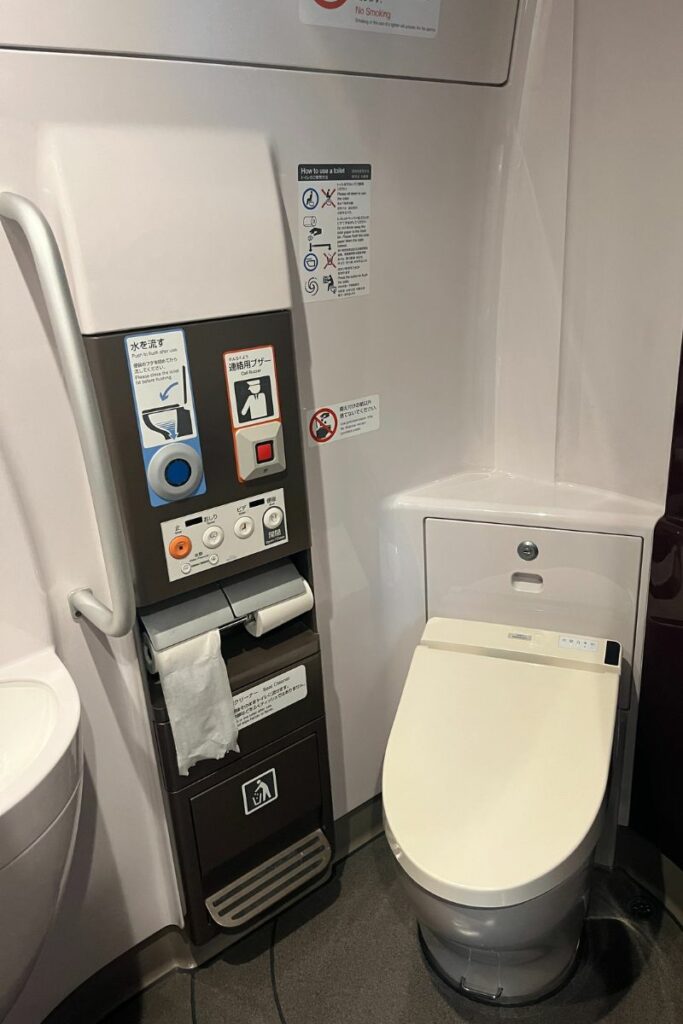 A view inside a restroom on the N700 Shinkansen. The image shows a modern toilet with an integrated bidet, control panel for flushing and bidet functions, a handlebar for stability, and a waste disposal area. The signage on the wall provides instructions for use and highlights the no-smoking policy.