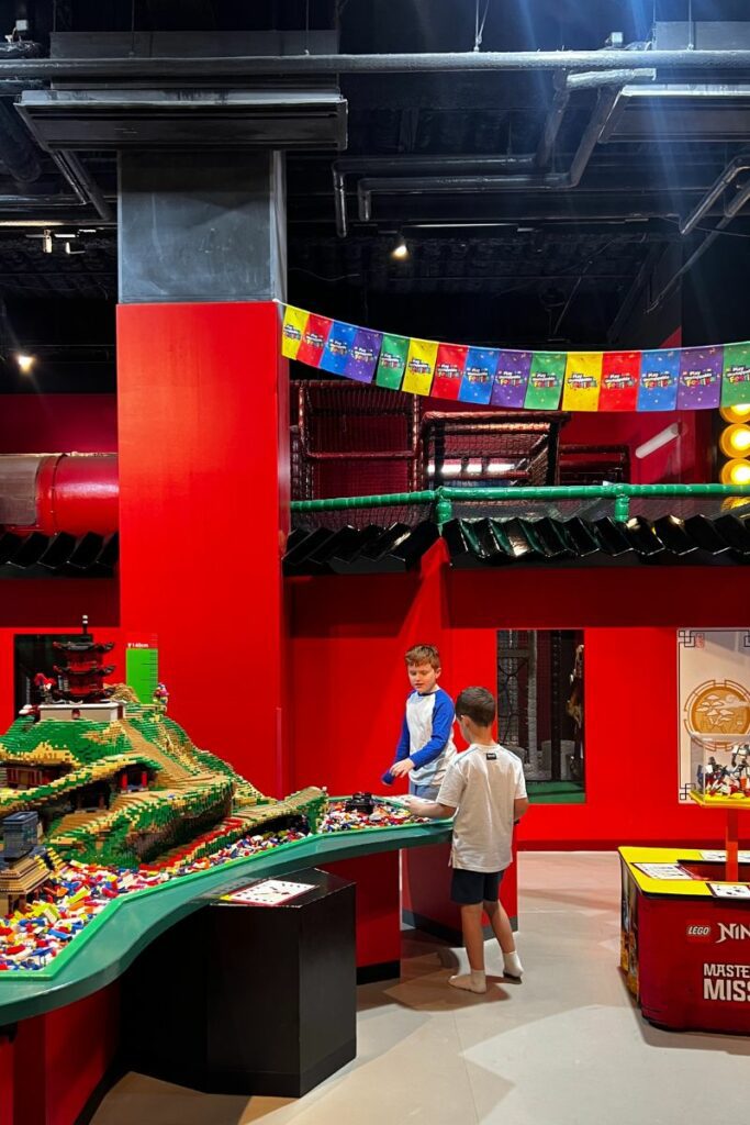 Two boys are playing in the Ninjago play area at LEGOLAND Discovery Center, surrounded by vibrant red walls and LEGO decorations. They are engaged in building with LEGO bricks on a table featuring a detailed Ninjago-themed landscape. Above them, colorful Ninjago banners are strung across the room.
