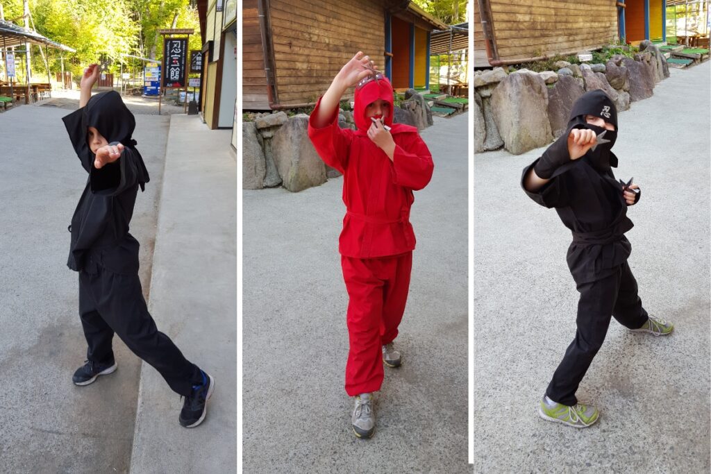 Three children dressed in ninja outfits strike poses with shurikens (ninja stars) in their hands. The first child, in a black outfit, poses with one arm raised. The second child, in a red outfit, holds a shuriken close to their face. The third child, also in a black outfit, holds a shuriken in a ready stance. The setting appears to be an outdoor area with wooden buildings and trees in the background.