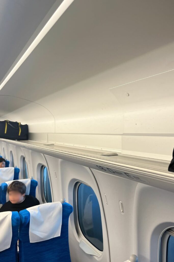 The overhead storage area in an ordinary class car of an N700 Shinkansen train is shown, with some luggage placed on the shelf. The seats below are blue with white headrest covers, and passengers are seated, some looking out the windows. The clean and spacious design of the storage area allows for ample luggage space above the seats.