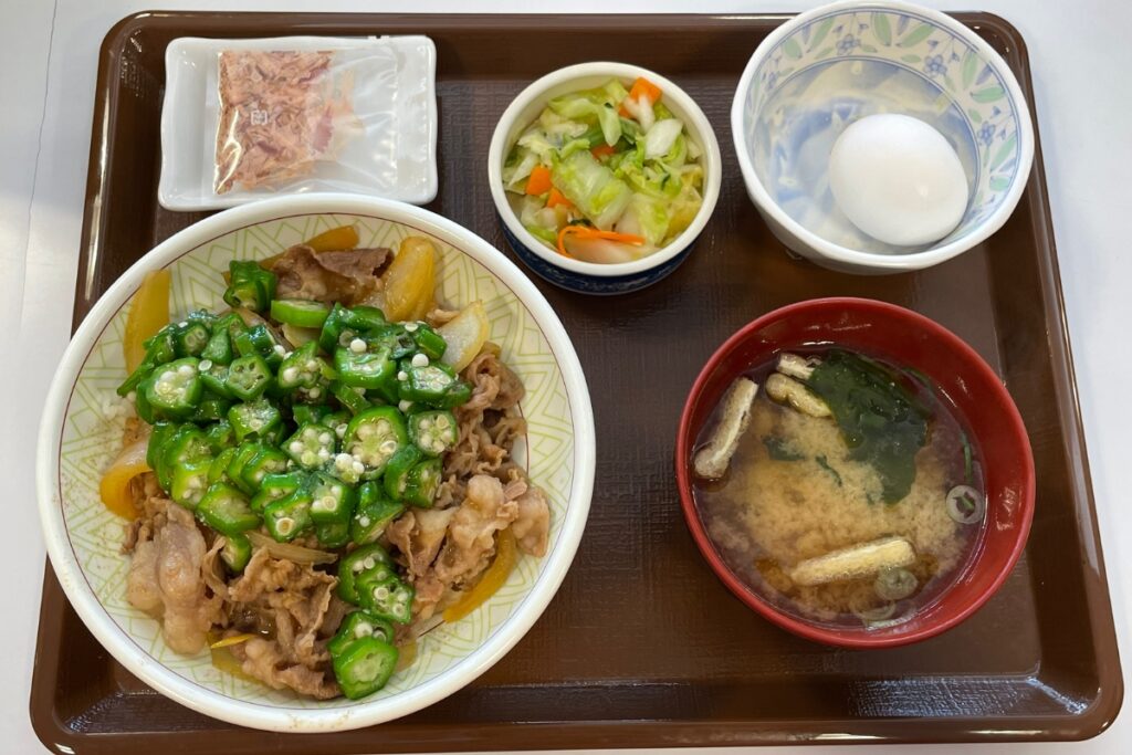 A tray with a Sukiya gyudon beef bowl topped with okra and onions, accompanied by a packet of dried bonito flakes, a small bowl of pickled vegetables, a soft-boiled egg, and a bowl of miso soup.
