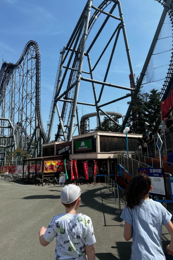 A boy and girl walking towards the Eejanaika roller coaster at Fuji-Q Highland. The massive coaster features complex loops and steep drops, constructed with grey steel beams. In the foreground, a Mos Burger fast food restaurant is visible under the coaster structure, with various promotional banners and signs around the entrance.