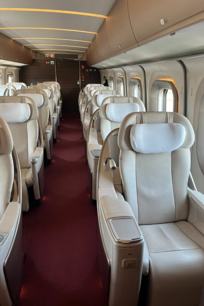 Interior of a Gran Class cabin on a Shinkansen train, featuring luxurious cream-colored leather seats with high backs and adjustable headrests. The cabin has a plush red carpet, subtle ambient lighting, and a quiet, refined atmosphere, emphasizing comfort and privacy.