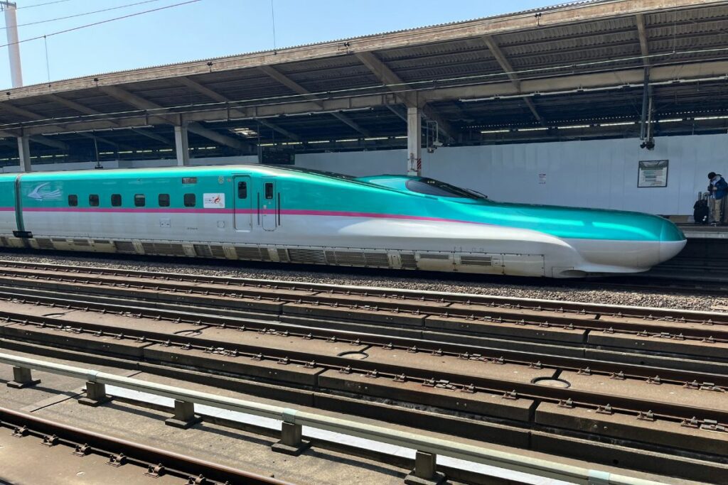 A side view of the iconic E5 Series Shinkansen, displayed at a train station under bright daylight. The high-speed train features a streamlined design with a distinctive green and pink stripe running along its white body. The train is on the tracks, set against the backdrop of the station's overhead structures, highlighting its sleek, modern engineering.