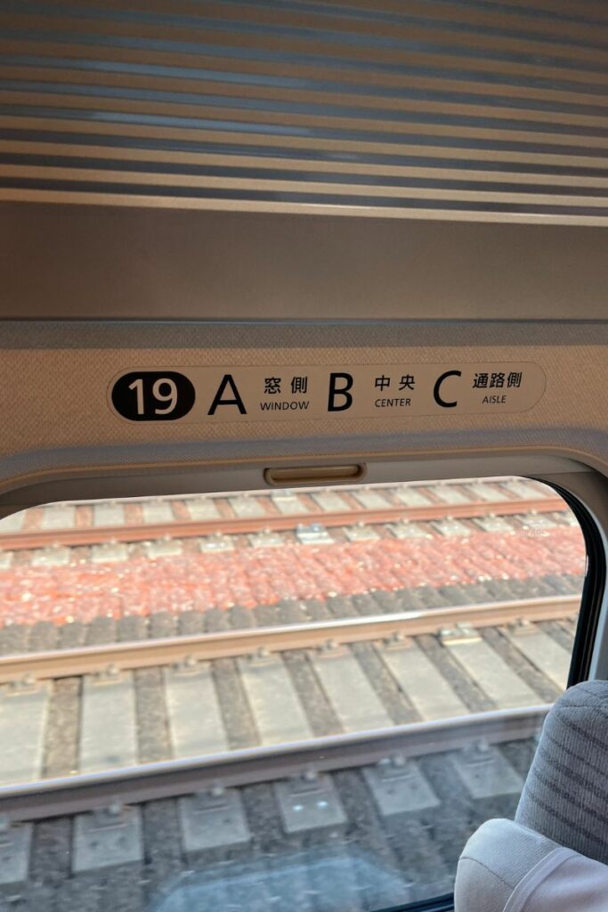 View from inside a train showing a window signage indicating seat numbers. The signage reads '19 A 窓 B 中 C 通路' corresponding to '19 A Window, B Center, C Aisle' in English, set against a backdrop of blurred train tracks seen through the window.