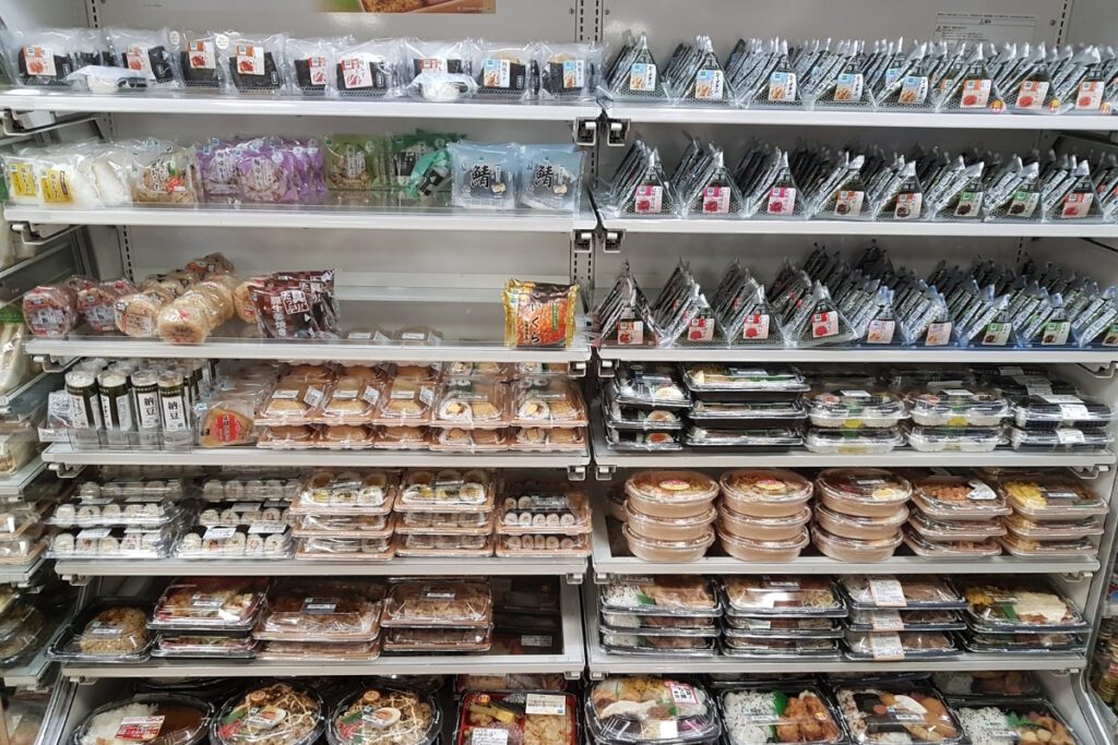 Shelves in a Japanese konbini (convenience store) stocked with a wide variety of packaged food items. The selection includes onigiri (rice balls), bento boxes, sandwiches, salads, and ready-to-eat meals, all neatly arranged and labeled. The display showcases the convenience and diversity of Japanese convenience store offerings.