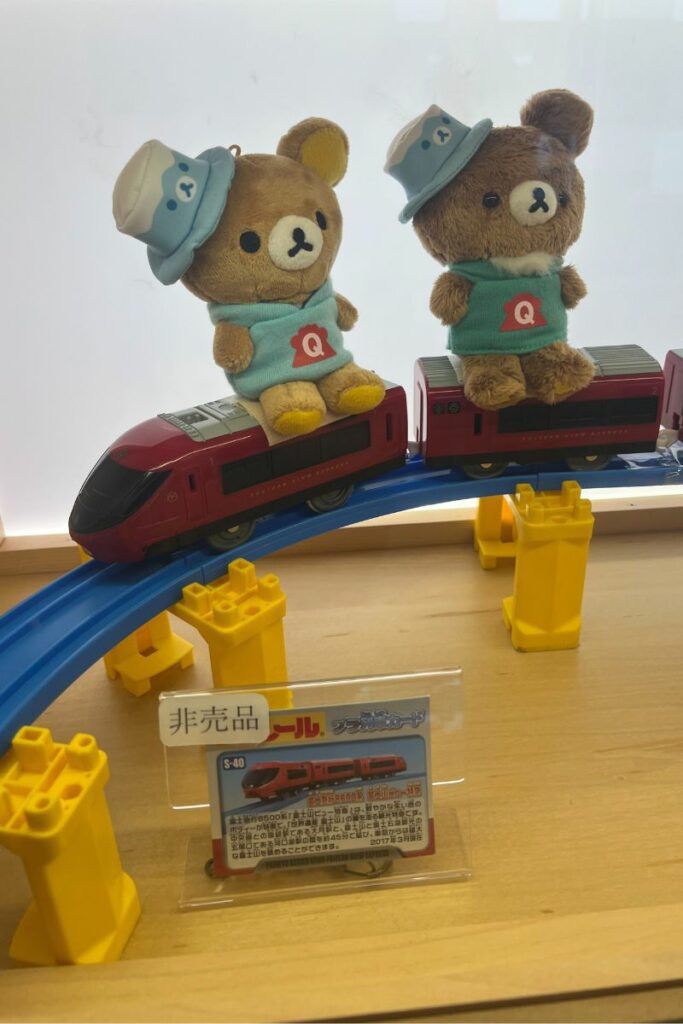 Two plush bear toys wearing blue hats and shirts with a "Q" logo, sitting on top of a miniature Fujisan View Express train model displayed on a blue track with yellow supports.