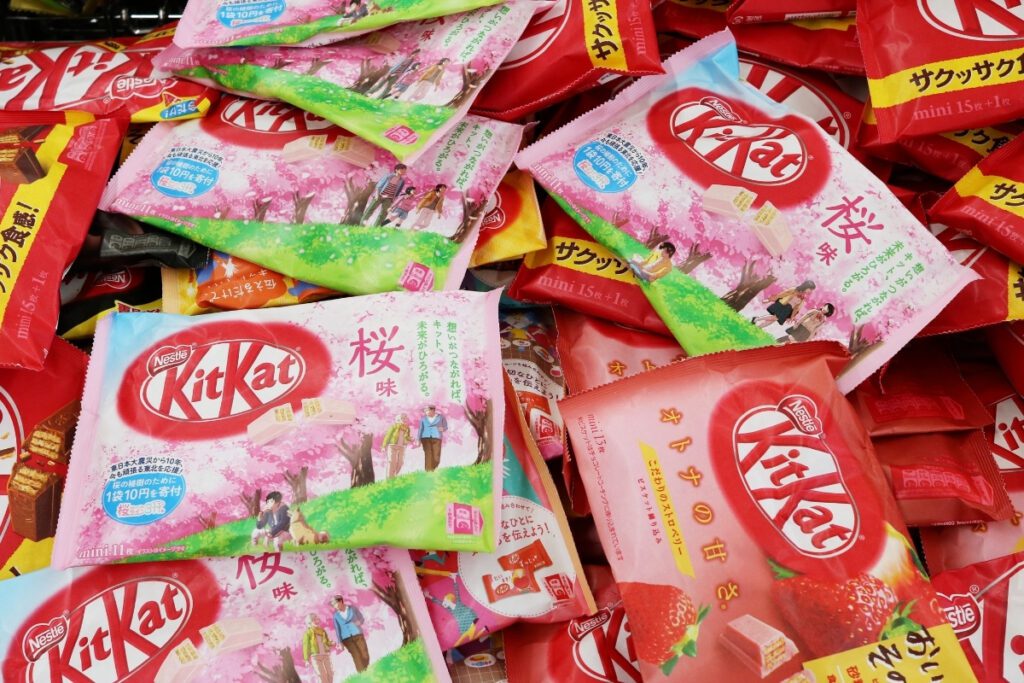 A colourful assortment of Kit Kat packages, showcasing a variety of flavors available in Japan. The packaging includes unique flavors such as cherry blossom (sakura) and strawberry, with vibrant designs featuring cherry blossoms and other Japanese-themed illustrations. The image highlights the diverse and creative range of Kit Kat options in Japan.