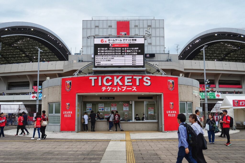 The ticket booth at Saitama Stadium, prominently labeled "TICKETS" with the Urawa Reds logo, where same-day tickets can be purchased. The electronic sign above displays information about upcoming home games in June. Fans in Urawa Reds jerseys are seen walking and standing in line to buy tickets.