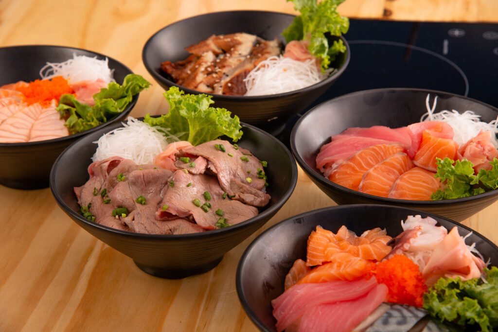 An assortment of donburi bowls, each featuring different toppings. The bowls include a variety of fresh sashimi such as salmon and tuna, cooked beef slices, and grilled fish, all garnished with lettuce, shredded daikon radish, and a dollop of roe. The bowls are arranged on a wooden table, showcasing the vibrant colors and diverse ingredients.