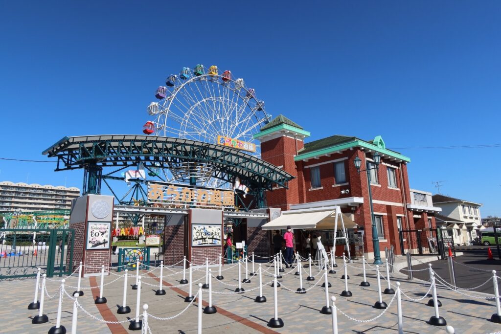 The entrance to Arakawa Amusement Park, featuring a decorative iron archway and a colorful Ferris wheel in the background. The red brick buildings on either side of the entrance add a classic touch to the vibrant park setting.