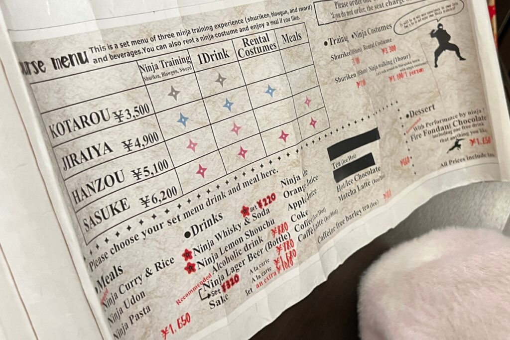 A close-up of a menu from the Ninja Experience Cafe in Harajuku, detailing various ninja training experiences, rental costumes, meals, and drinks. The options include packages named Kotarou, Jiraiya, Hanzou, and Sasuke, each with different combinations of training, drinks, and meals. The menu also lists specific food and drink items such as Ninja Curry & Rice, Ninja Udon, Ninja Pasta, Ninja Whisky & Soda, and Ninja Lager Beer.