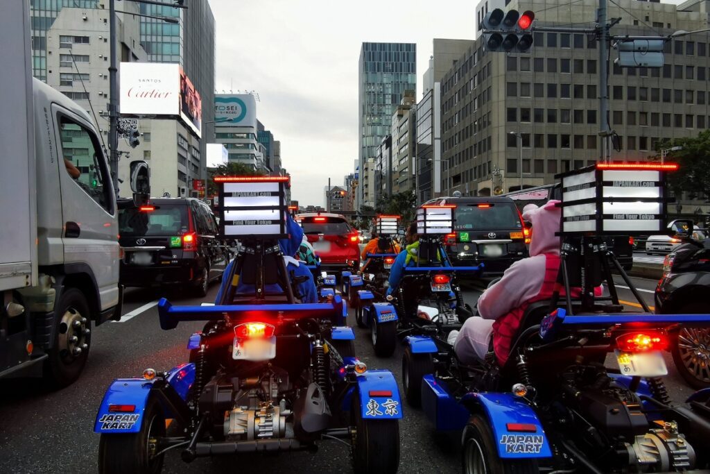 A group of people driving go-karts through the busy streets of Tokyo, dressed in various costumes. The go-karts are blue with "Japan Kart" decals, and the drivers are waiting at a red traffic light surrounded by cars and trucks. Skyscrapers and commercial buildings line the street, with prominent signs for brands like Cartier visible in the background.