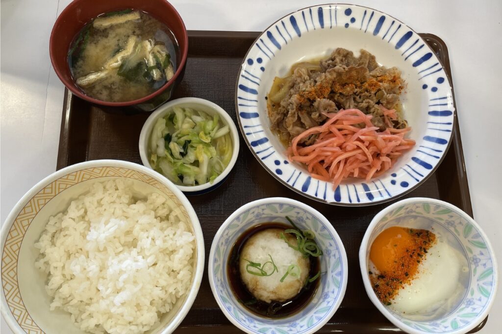 A meal tray at Sukiya with several dishes. The tray includes a bowl of rice, a bowl of miso soup with tofu and green onions, a dish of beef with pickled ginger, a bowl of grated radish with soy sauce and green onions, and a bowl with a soft-boiled egg topped with seasoning.