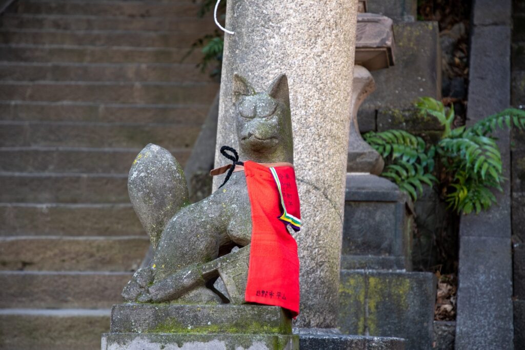 Stone fox statue at Oji Inari Shrine, adorned with a red bib and positioned next to a stone pillar with stairs in the background.