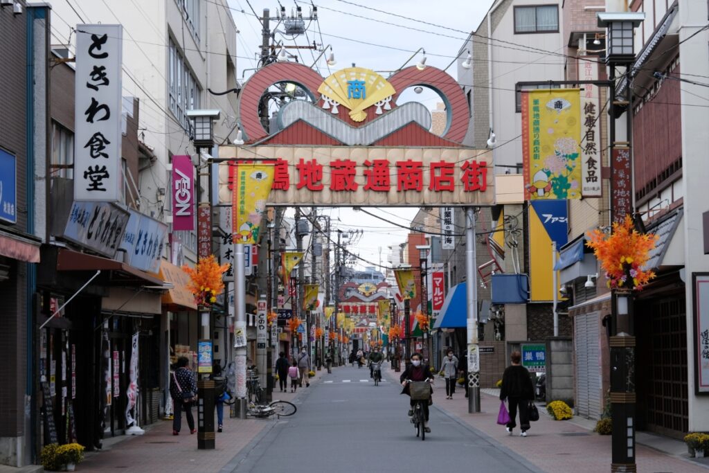 Sugamo Jizodori Shopping Street, featuring colorful signs and banners, various shops lining the street, and a few pedestrians and cyclists. The archway sign is prominently displayed in the center.