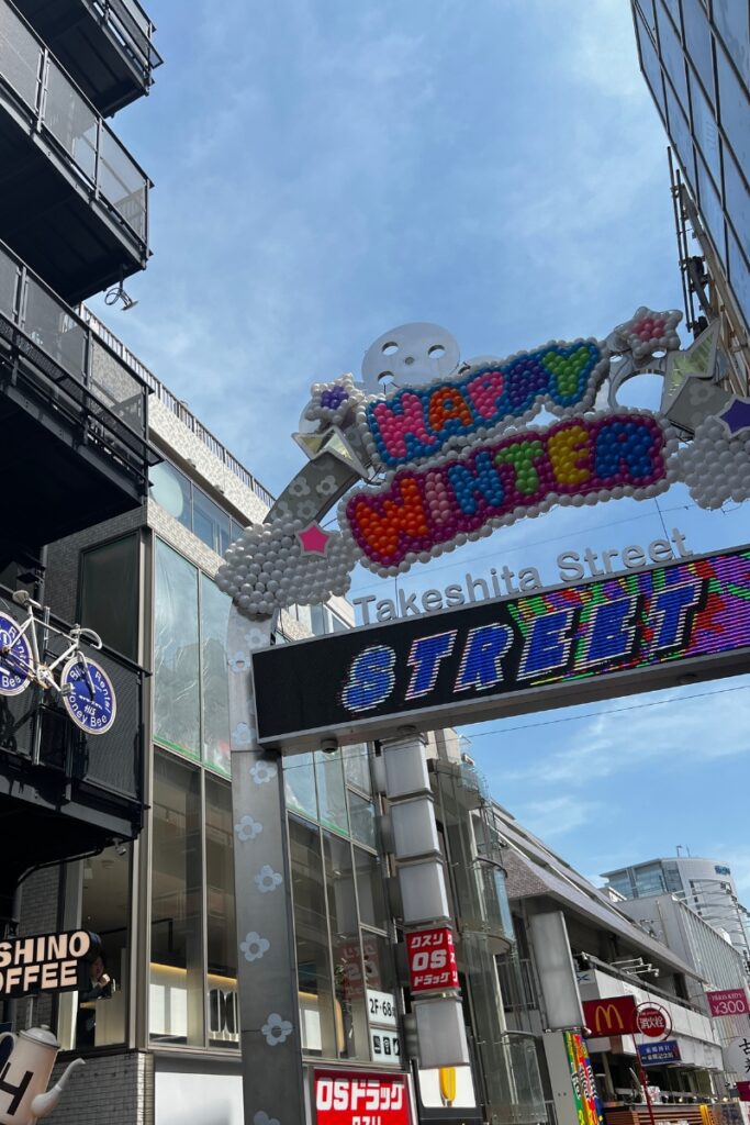 The colorful entrance sign to Takeshita Street in Harajuku, Tokyo, decorated with balloon art that reads "Happy Winter." The sign is surrounded by buildings with various shops, including a coffee shop and a McDonald's, under a clear blue sky.