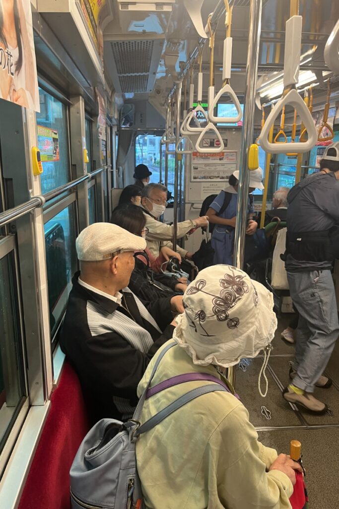 Passengers, including elderly people, seated inside a crowded Tokyo Sakura Tram, with standing passengers holding onto the handrails. The atmosphere is calm, with everyone quietly commuting.