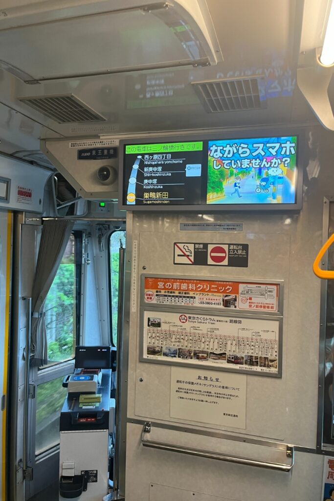 Interior of a Tokyo Sakura Tram showing the route map and next stops on a digital display behind the driver's seat. The display indicates upcoming stops: Nishigahara-yonchome, Shinkoshinzuka, Koshiuzuka, and Sugamo-Shinden. An advertisement and no-smoking sign are also visible.