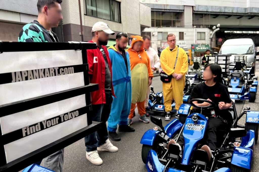 A group of people in colorful costumes standing next to blue go-karts, receiving a demonstration from a staff member on how to drive the go-karts. The staff member is sitting in one of the go-karts, while the group listens attentively. The go-karts are branded with "Japan Kart" and "Find Your Tokyo" decals.