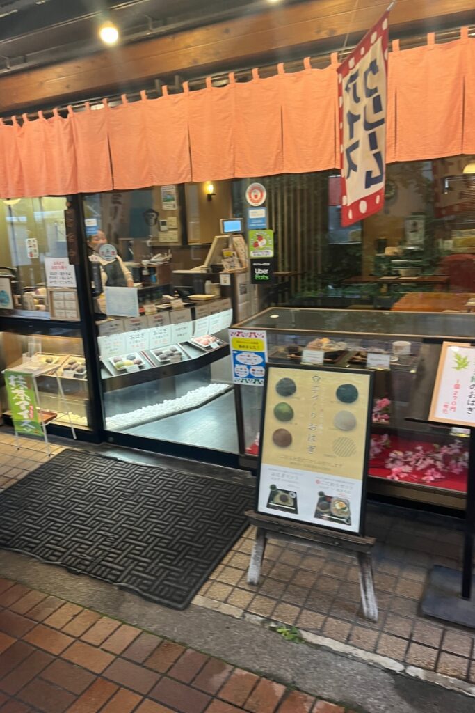 Exterior view of Sweets Shop Ippukutei at Koshinzuka Station, featuring a traditional Japanese curtain, display cases with various sweets, and a signboard with menu items. A person is visible inside the shop near the counter.