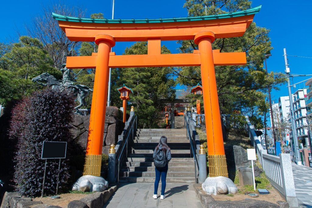 A large orange torii gate marks the entrance to Ana-Hachimangu Shrine. A woman stands at the base of the steps leading up to the shrine, which is surrounded by greenery and traditional lanterns. The sky is clear and blue, adding to the serene atmosphere.