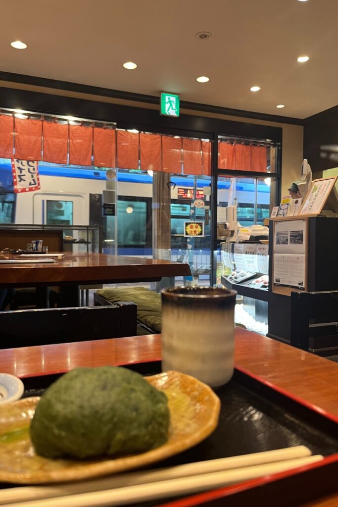 Interior view of Sweets Shop Ippukutei at Koshinzuka Station, showing a table with a plate of ohagi (a Japanese sweet rice ball) and a cup of matcha. The tram is visible through the window, reflecting the shop's proximity to the station platform.