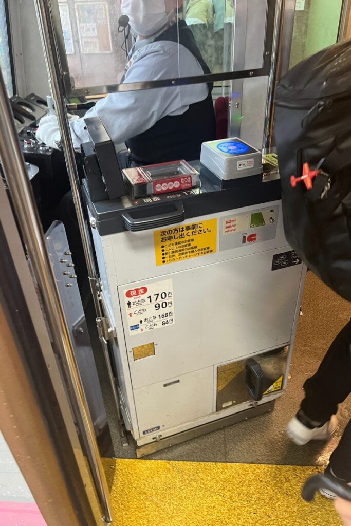 Close-up of a fare payment machine inside a Tokyo Sakura Tram, where passengers can pay with cash or IC cards like Suica. A tram conductor wearing a mask is visible behind the machine. The fare for adults is 170 yen and 90 yen for children.