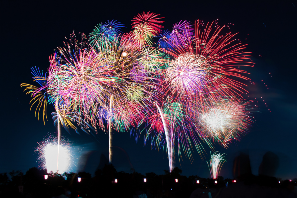 A vibrant fireworks display in the night sky with multiple bursts of color, including red, green, blue, and purple, creating a stunning array above a dark silhouette of trees. The bright, multi-colored explosions fill the sky with dazzling light, illuminating the scene with a festive energy.