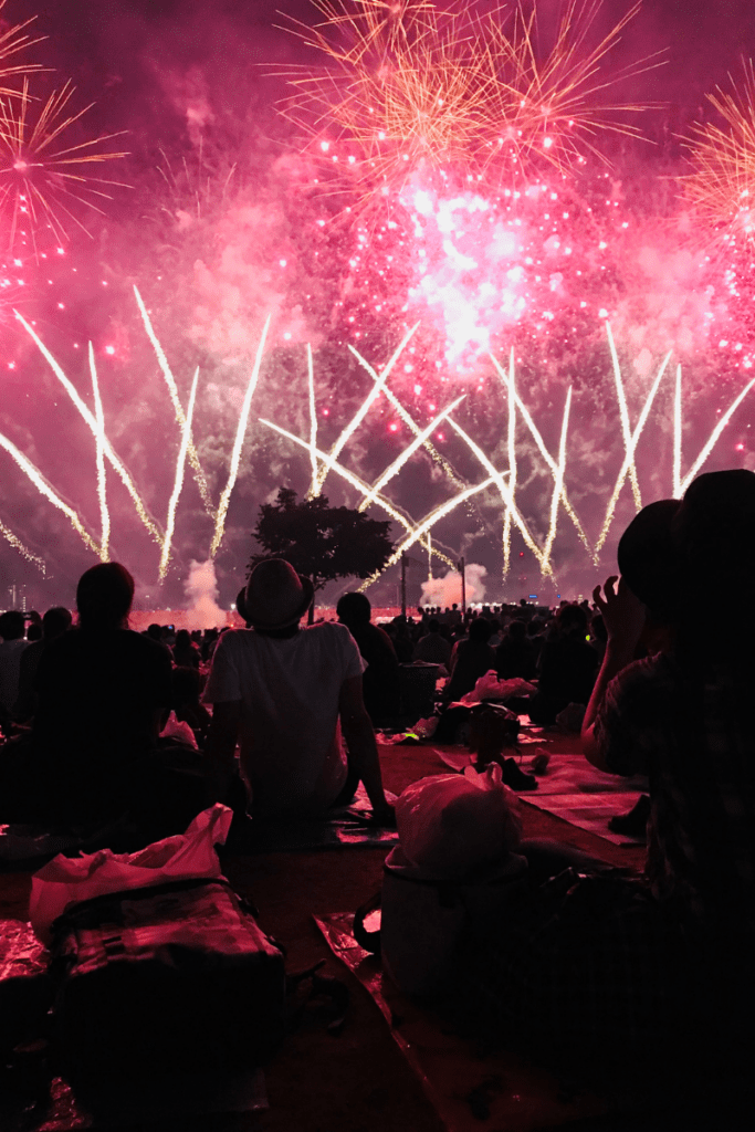 The image captures a vibrant fireworks display lighting up the night sky with brilliant pink, red, and gold colors. The fireworks form a stunning array of patterns, including starbursts and crisscrossing trails. Silhouetted against this dazzling backdrop are people sitting on blankets, watching the show in awe, creating a lively and festive atmosphere.