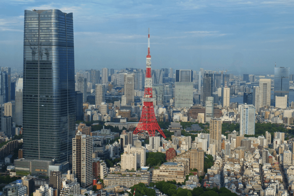 The image shows a stunning view of Tokyo's skyline, with the iconic red and white Tokyo Tower standing prominently in the center. Surrounding the tower are a mix of modern skyscrapers and smaller buildings, including the sleek Roppongi Hills Mori Tower to the left. The expansive urban landscape stretches into the distance, with a blue sky and light clouds overhead, capturing the vastness and density of Tokyo.