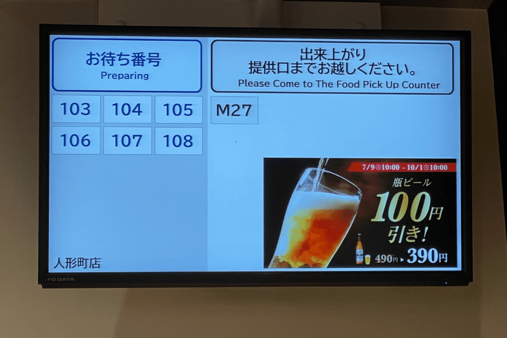 This image shows a digital order screen at a Matsuya restaurant. The screen displays order numbers under a "Preparing" section, including 103, 104, 105, 106, 107, and 108. One completed order, labeled M27, is ready for pickup. The screen also features a promotion for bottled beer, offering a discount of ¥100, reducing the price from ¥490 to ¥390. The text above instructs customers to pick up their orders at the food counter when ready.