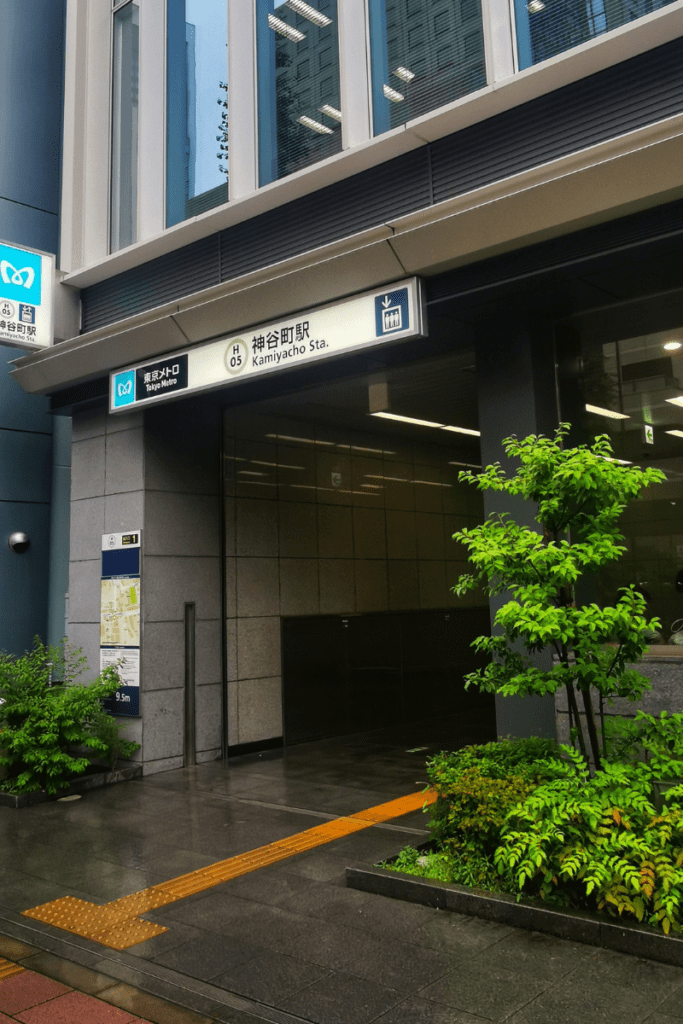 The image shows the entrance to Kamiyacho Station, part of the Tokyo Metro. The station signage is prominently displayed above the entrance, labeled with the station code "H05" and the station name in both Japanese and English. The entrance is modern and sleek, with clean stone walls and glass windows. A small patch of greenery, including shrubs and a small tree, decorates the foreground next to the walkway, and a tactile yellow path for the visually impaired leads into the station. The surrounding area appears clean and well-maintained.