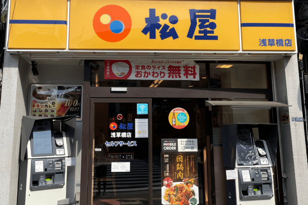 This image shows the exterior of a Matsuya restaurant in Japan, with its bright yellow and blue signage displaying the restaurant's name in Japanese. The entrance features large windows and two self-service kiosks for ordering and payment, one on each side of the door. Promotional signs advertise new menu items, including chicken curry for ¥480 and iced drinks for ¥100 off. A banner above the door highlights free rice refills with certain meals.